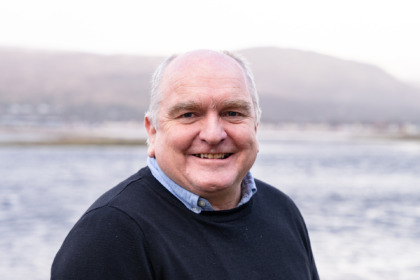 Frazer Coupland, Chief Executive of West Highland Chamber, stands by a loch, with mountains behind him. He is wearing a woolly jumper and smiling at the camera.